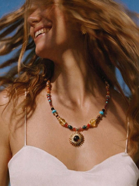 Femme souriante au soleil portant le collier DUNE DORÉE, un ras de cou bohème chic avec pendentif soleil en œil de tigre.
