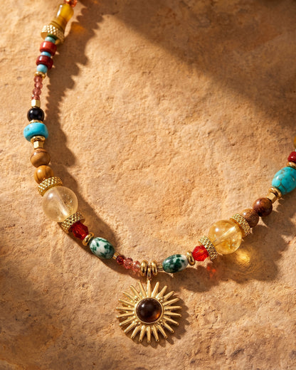 Gros plan sur le pendentif soleil et les perles en pierres naturelles du collier DUNE DORÉE, posé sur le sable chaud.
